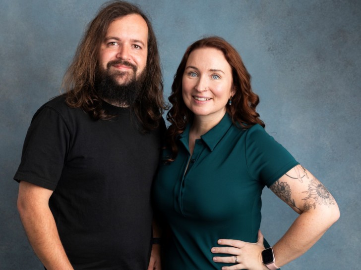 A woman with long auburn hair and a man with long brown hair stand side by side against a plain background. They are each facing the camera and smiling.