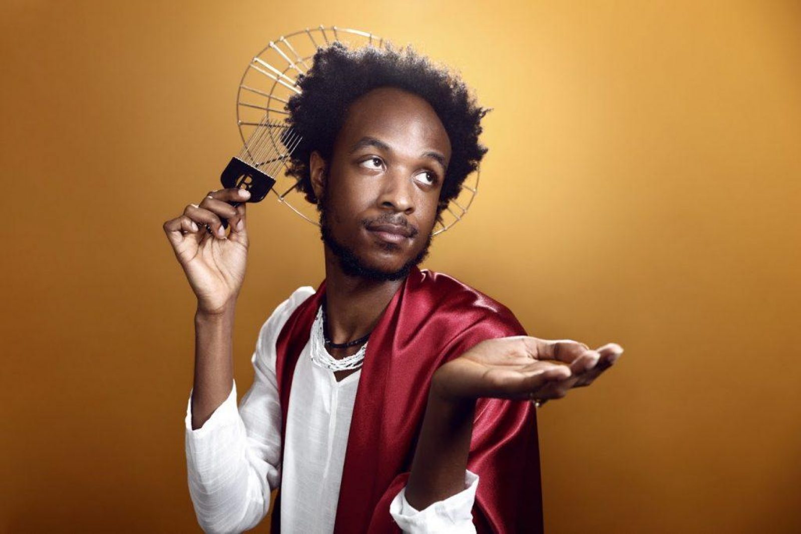 Jamal Gerald in front of a gold background, wearing a white linen shirt, a silk red cape and a metal halo, he has an afro comb in one hand and holding the other hand palm upwards in front of him.