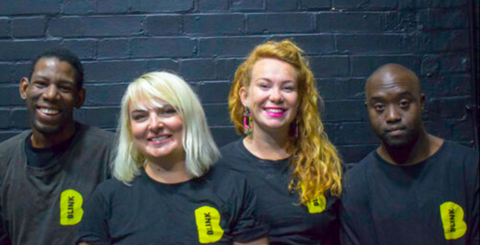 4 members of blink Dance theatre looking at the camera, stood in front of a black wall, wearing black T-shirts with the company logo on.
