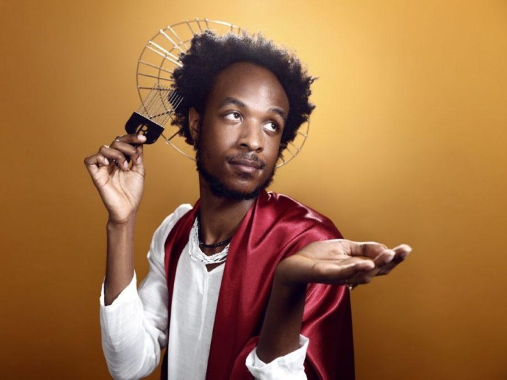 Jamal Gerald in front of a gold background, wearing a white linen shirt, a silk red cape and a metal halo, he has an afro comb in one hand and holding the other hand palm upwards in front of him.