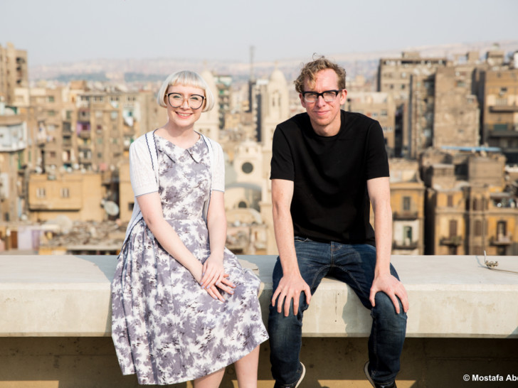 Andy and Beckie sit next to each other on a wall. Behind them is a cityscape full of multi-story buildings.