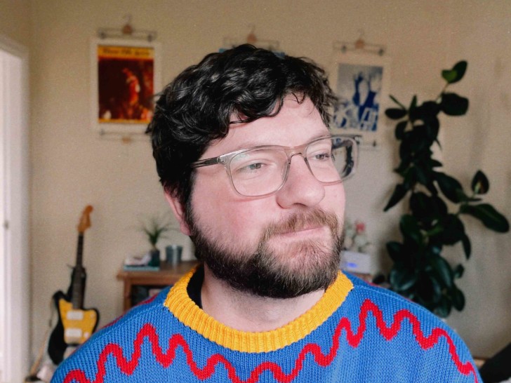 A man with short dark hair wearing glasses and a colourful blue, red and yellow jumper sits in a room looking to one side and smiling. Behind them is large plant and a guitar.