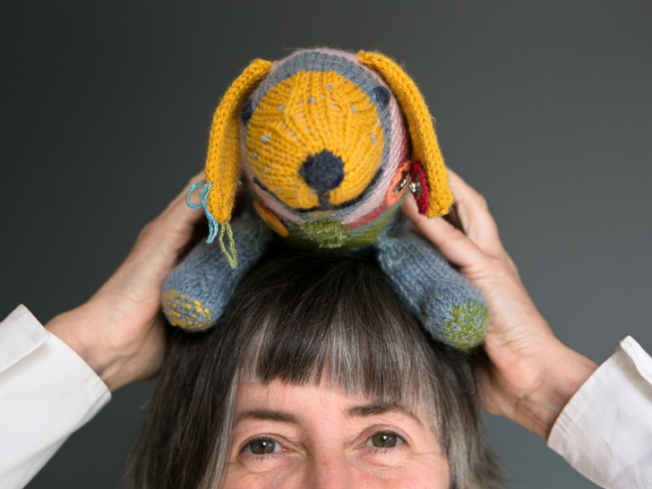 Deirdre Nelson photographed up close from the eyes up holding a knitted dog on her head.