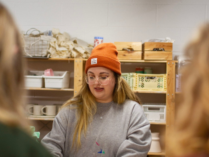 Emma is hosting a workshop in which she is working with clay, she wears an orange beanie and a grey jumper.