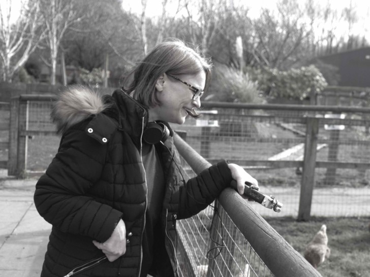 This black and white photo shows a person with light, bob-cut hair, who is wearing glasses and a black coat with a fur trim. The person is learning on a fence into a chicken pen, looking into the pen and holding a zoom recorder in their left hand.