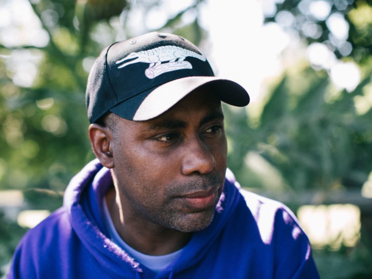 Harun Morison is photographed looking to the right of the camera, wearing a black baseball cap with a crocodile on it, there are trees in the background.