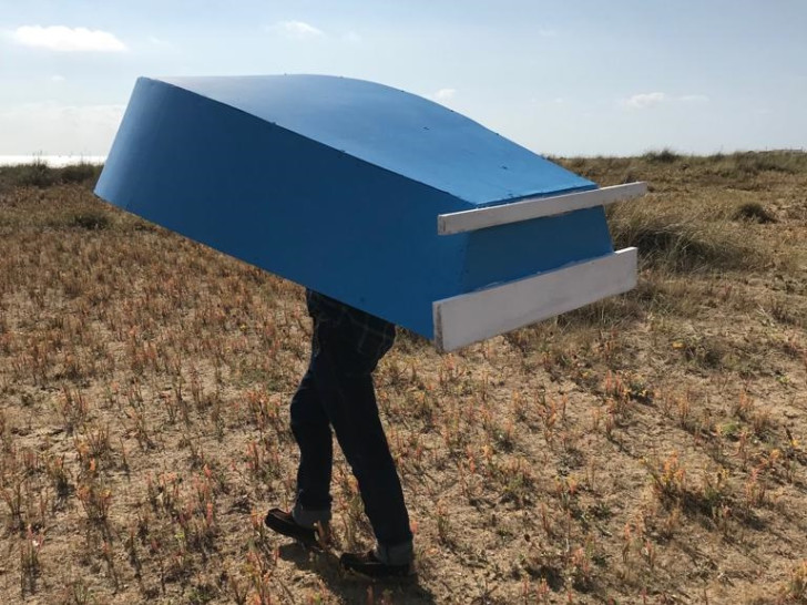 Mark Storor walking uphill on a field whilst carrying a large blue wooden object above his head.