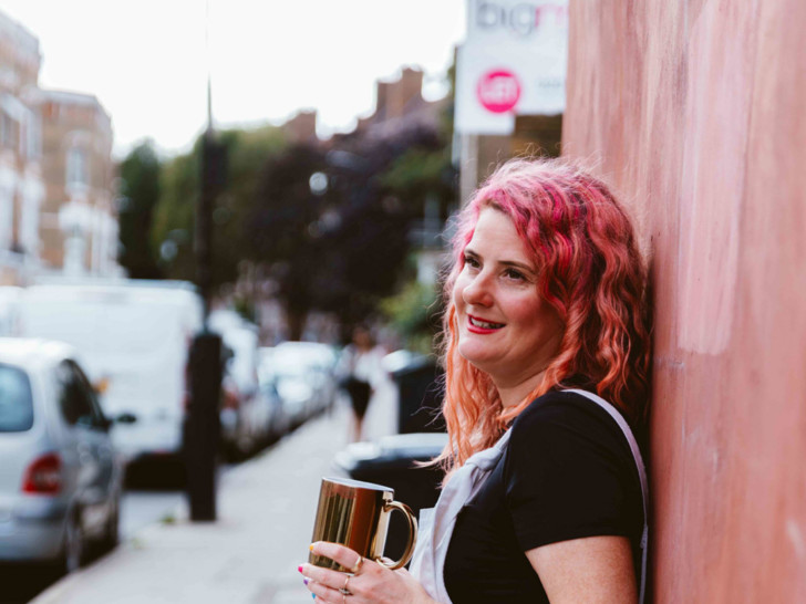 Kelly is stood leaning against a wall, they have pink hair and she is holding a gold mug in her hand, wearing white dungarees and a black t shirt.
