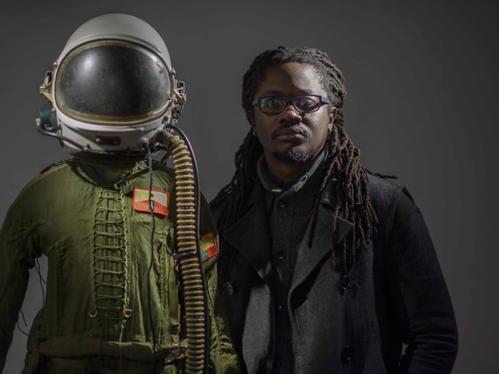 A black man with long dreadlocks and wearing glasses, a dark jacket and wearing glasses stands next to a figure wearing a vintage white space helmet and wearing a green top that is laced together down the front.