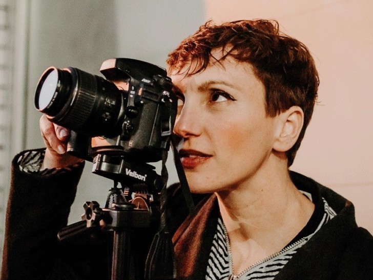 A white woman with cropped red hair looks upwards through the lens of a DLSR camera