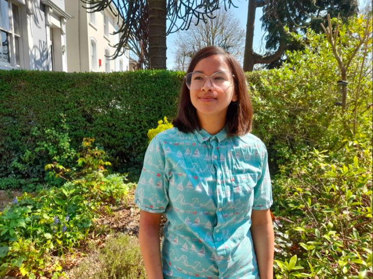Pippa Sterk is photographed in a garden on a sunny day. They wear a turquoise shirt and a pair of spectacles with round rims.