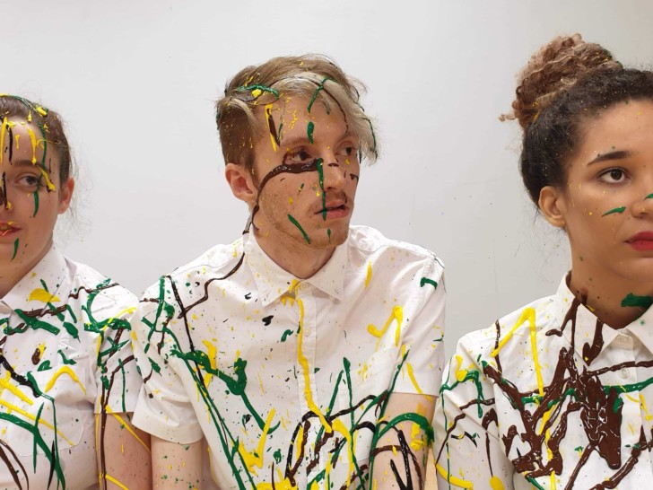 The three members of the 'Year of the Rat Collective' sit next to each other in a line. The are all wearing white shirts and are splattered with green, yellow and back paint