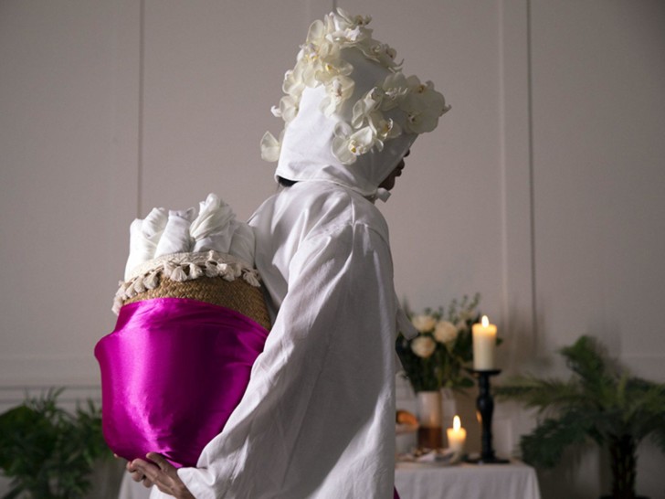 A person wearing a white cotton gown, with a white cotton hood adorned with white flowers carries a basket wrapped in pink satin fabric on their back. The basket is full of white fabric rolled up. Behind them a table with candles, a large fruit bowl and white flowers is set up like as altar.