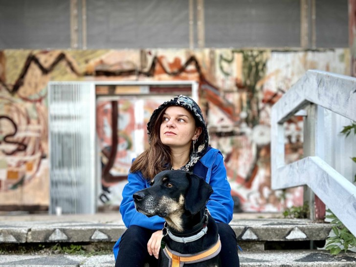 Zorka Woolly sitting on some concrete stairs in front of an industrial building covered in graffiti, with a dog sat in between her legs.