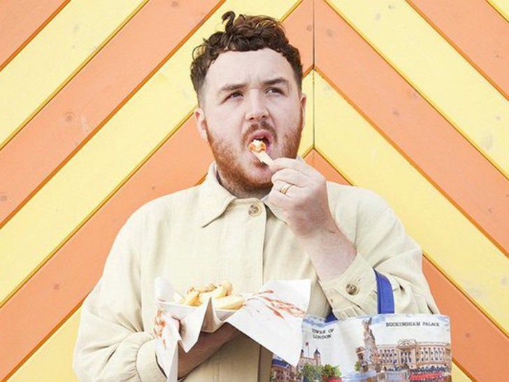 Scottee is photographed in front of a background that is yellow and orange planks of wood, eating chips looking upwards with a confused expression.