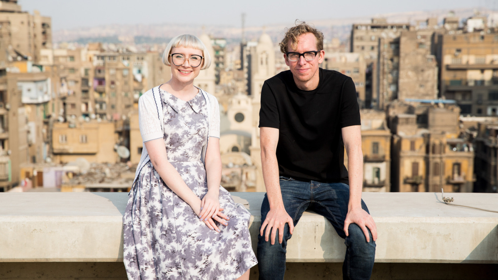 Andy and Beckie sit next to each other on a wall. Behind them is a cityscape full of multi-story buildings.