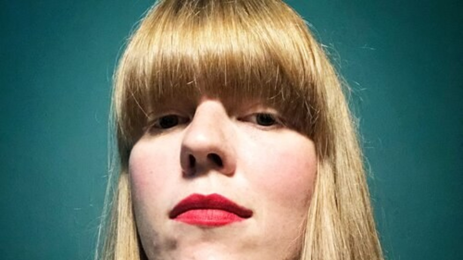 Fox Irving is looking down at the camera, she is stood in front of a dark green Backdrop, and has medium length straight strawberry blonde hair with a fringe. She is wearing red lipstick.