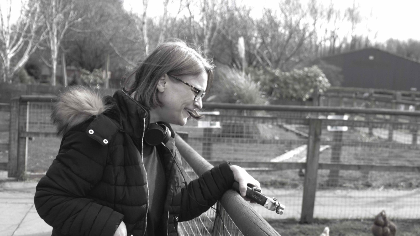This black and white photo shows a person with light, bob-cut hair, who is wearing glasses and a black coat with a fur trim. The person is learning on a fence into a chicken pen, looking into the pen and holding a zoom recorder in their left hand.