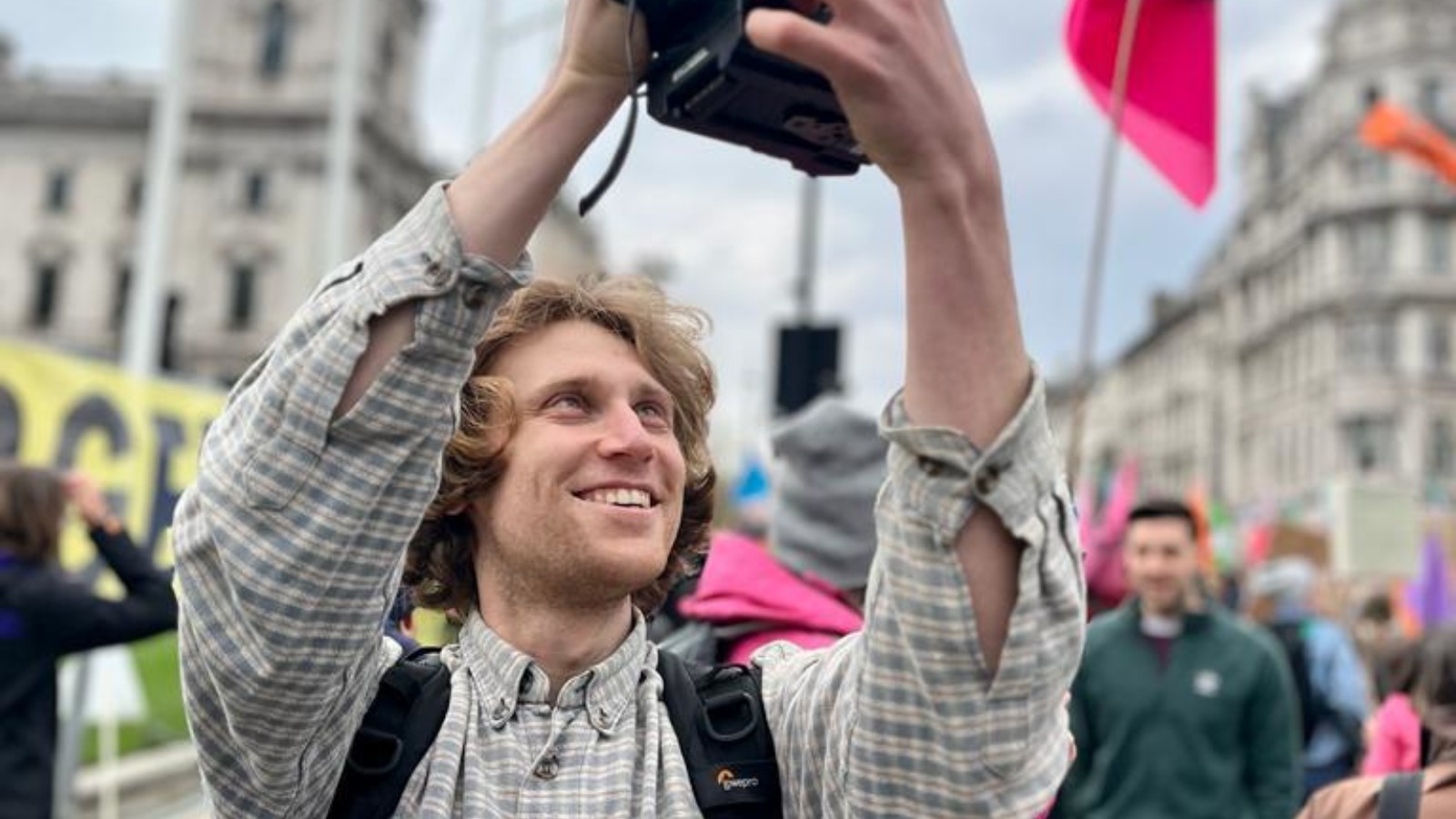 Rich Felgate is photographed filming at a march through a city, he is smiling and wearing a checkered shirt.