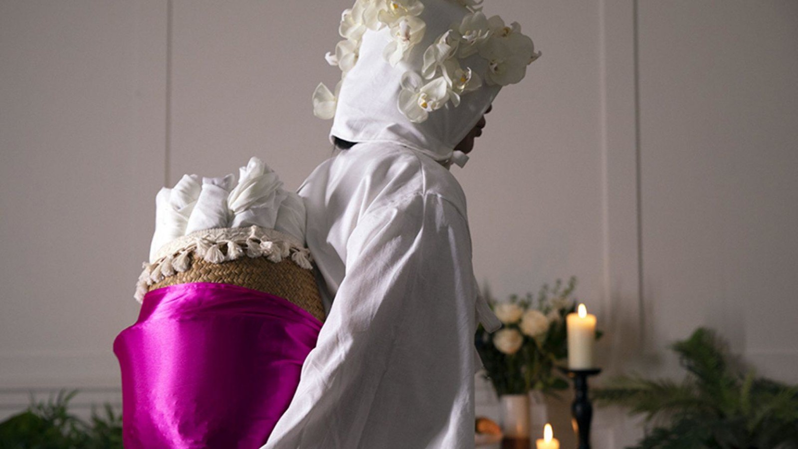 A person wearing a white cotton gown, with a white cotton hood adorned with white flowers carries a basket wrapped in pink satin fabric on their back. The basket is full of white fabric rolled up. Behind them a table with candles, a large fruit bowl and white flowers is set up like as altar.