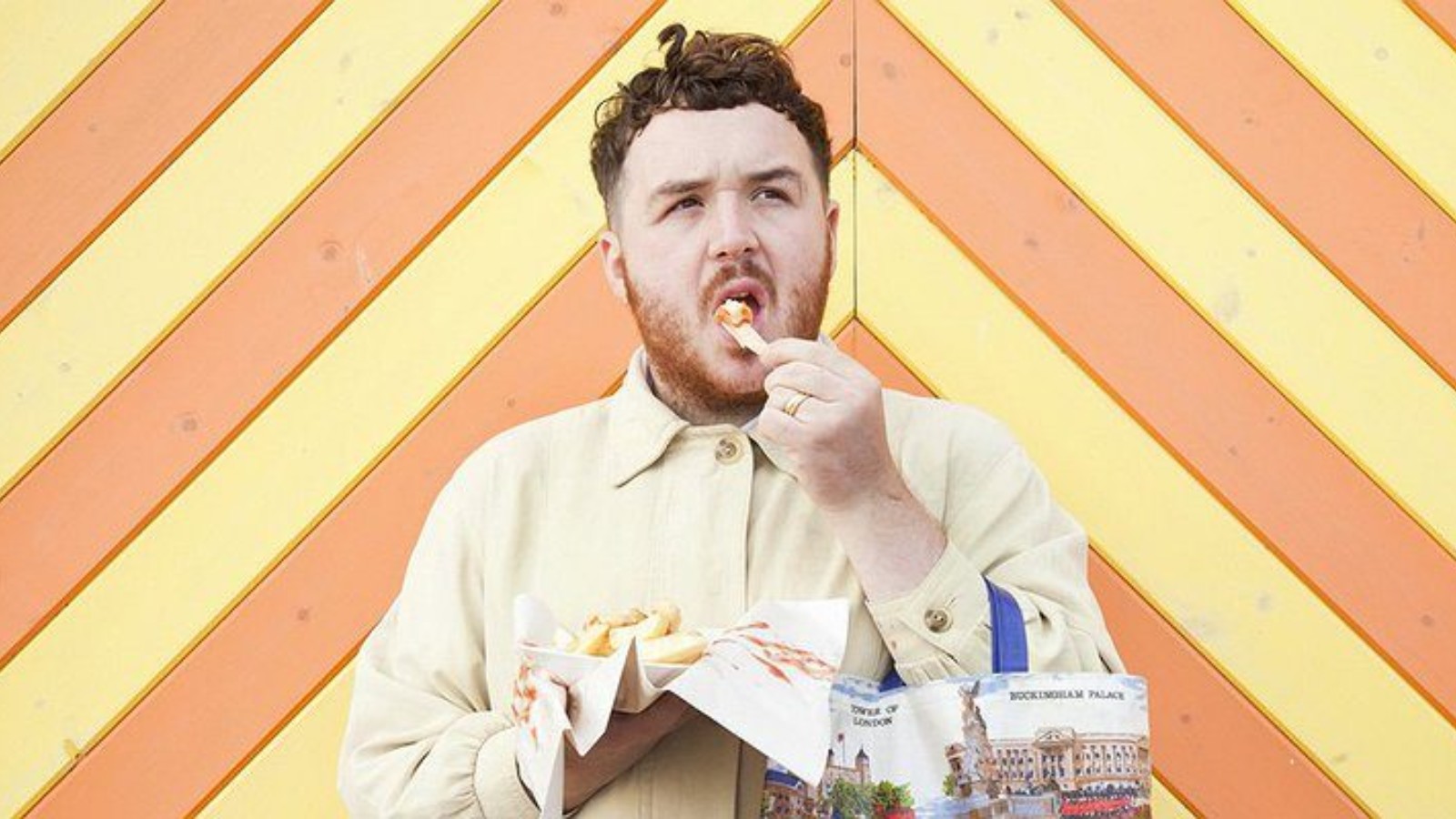 Scottee is photographed in front of a background that is yellow and orange planks of wood, eating chips looking upwards with a confused expression.