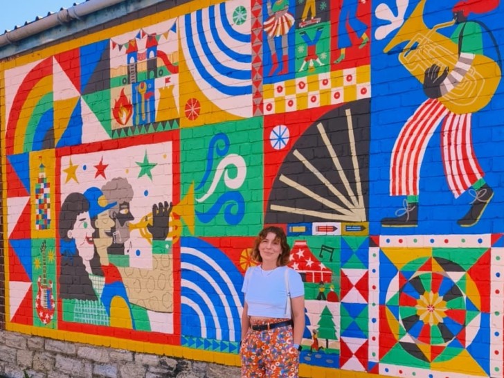 A woman with red hair stands smiling in front of a large colourful mural.