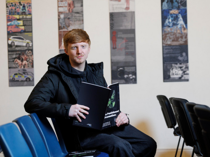 A man with red hair, a short moustache and beard sits in a row of empty blue chairs holding a booklet open in their lap.