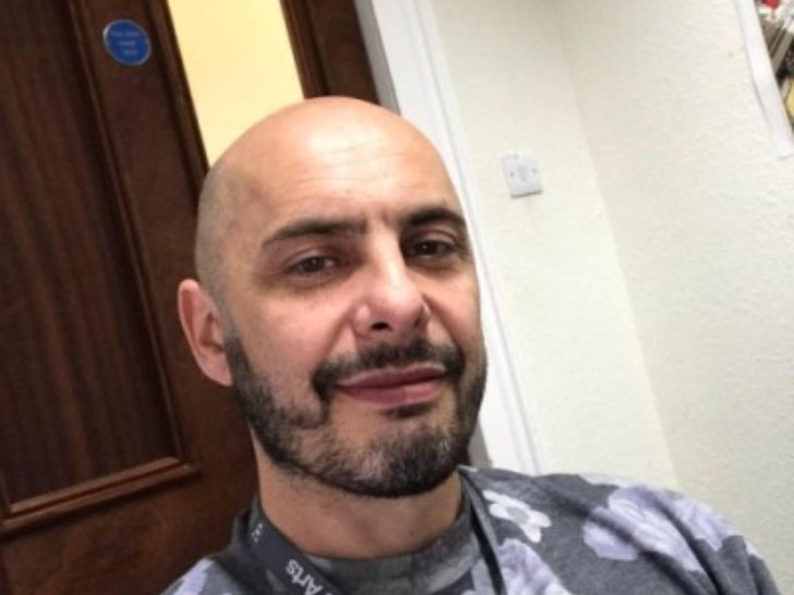 A man with a short dark beard and moustache and wearing a dark lanyard that reads 'School of the Arts' looks at the camera.