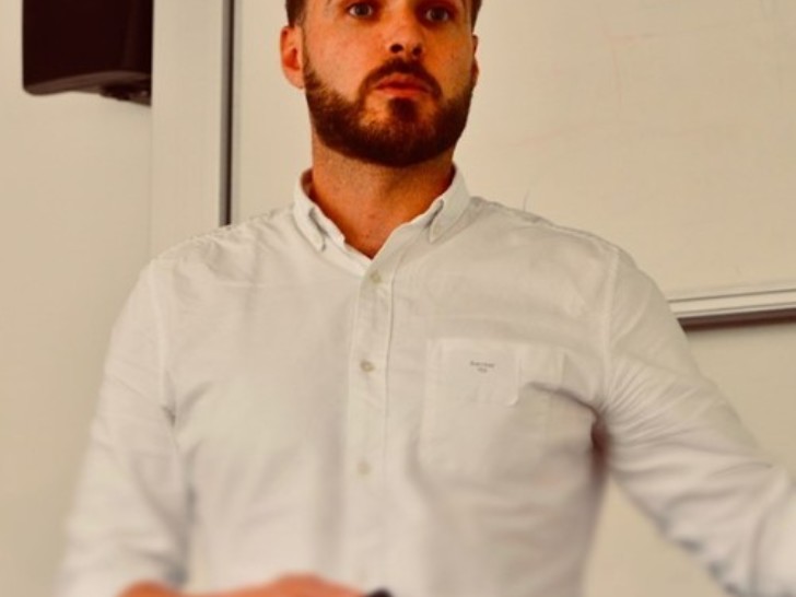 A man with dark brown hair and facial hair wears a white shirt and is talking in front of a white background