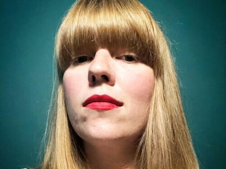 Fox Irving is looking down at the camera, they are stood in front of a dark green Backdrop, and have medium length straight ginger hair with a fringe.