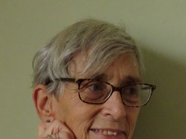 A portrait of a white women on a green background. The woman has short grey hair, is wearing dark rimmed glasses, a blue and white patterned shirt and is smiling slightly.