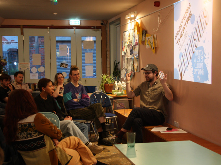 Photograph of a man wearing a cap and brown shirt, sat in a cafe with low lighting. The man is giving a presentation to a small audience, with an image projected on the wall behind him.
