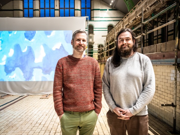 Juneau Projects stand in front of an abstract blue projection, smiling a the camera.
