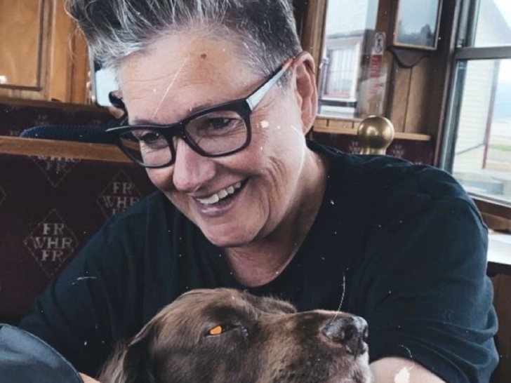 A woman with cropped hair and dark glasses is smiling and looking in their bag. They have a brown dog's face resting on their arm.