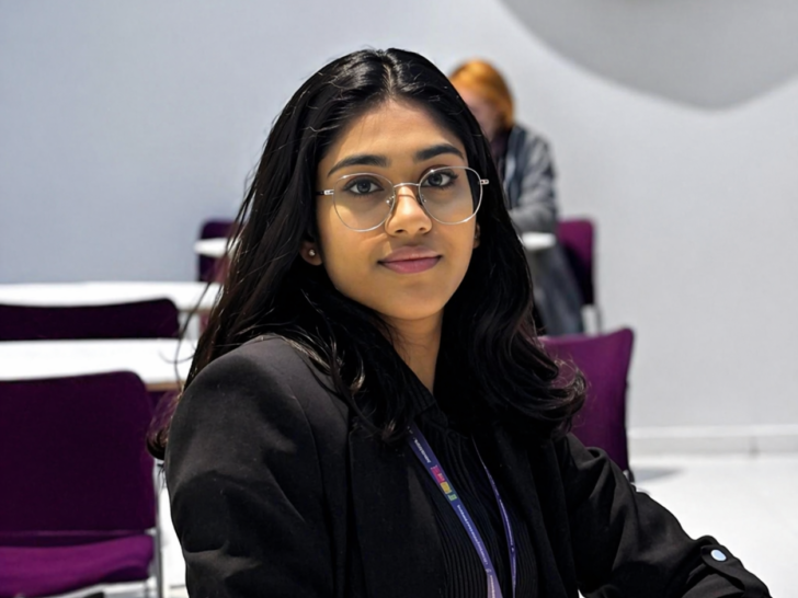 A woman with dark hair wearing a black blazer, glasses and a purple lanyard sits in a large white room.