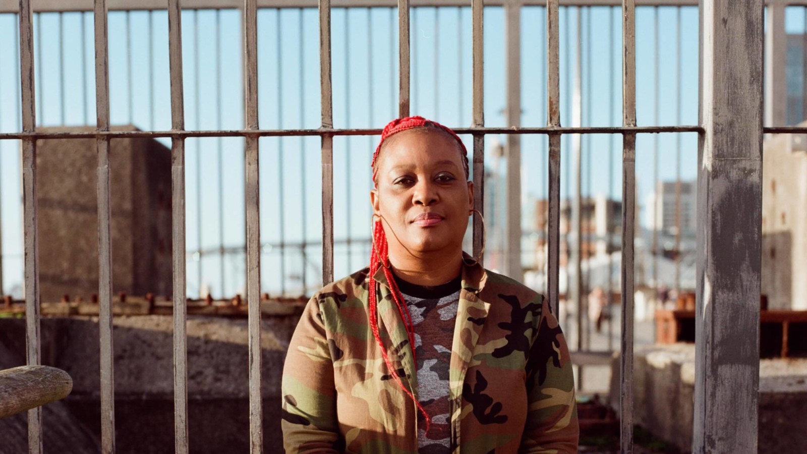 A black woman with red braids sits on a low stone wall, with railings behind her.