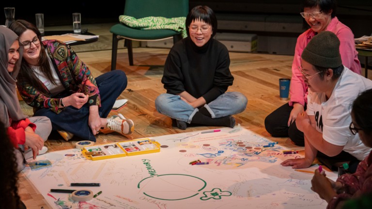 A group of people sit and kneel on a floor around a large piece of paper covered in colourful markings. They are laughing and looking at the paper.