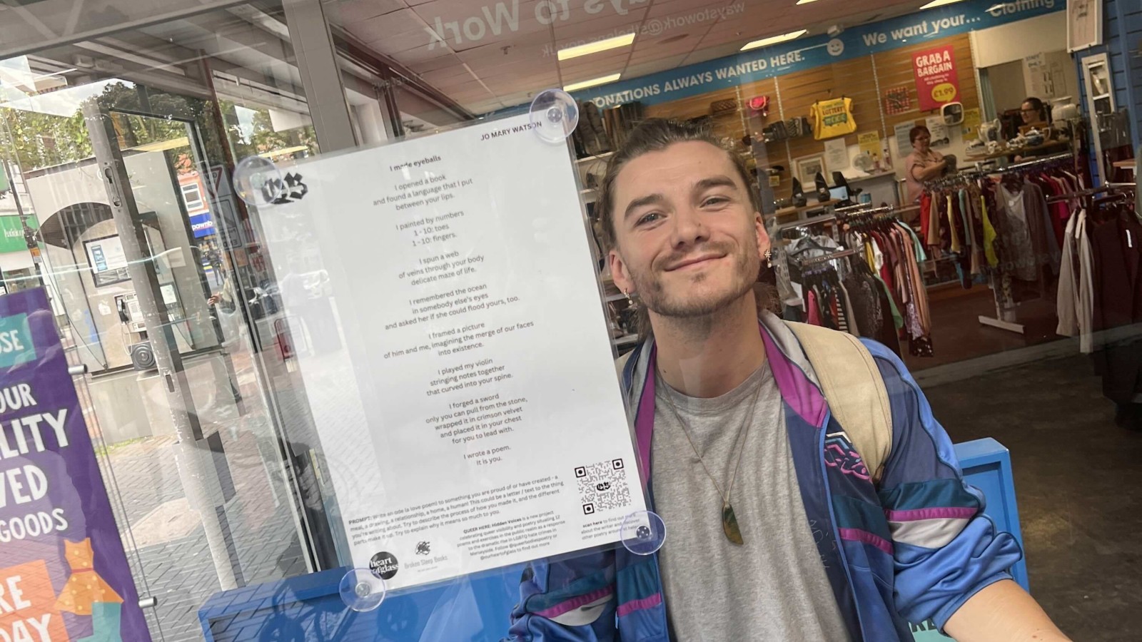 A poem in the window of a shop, with a long haired smiling person smiling through the glass