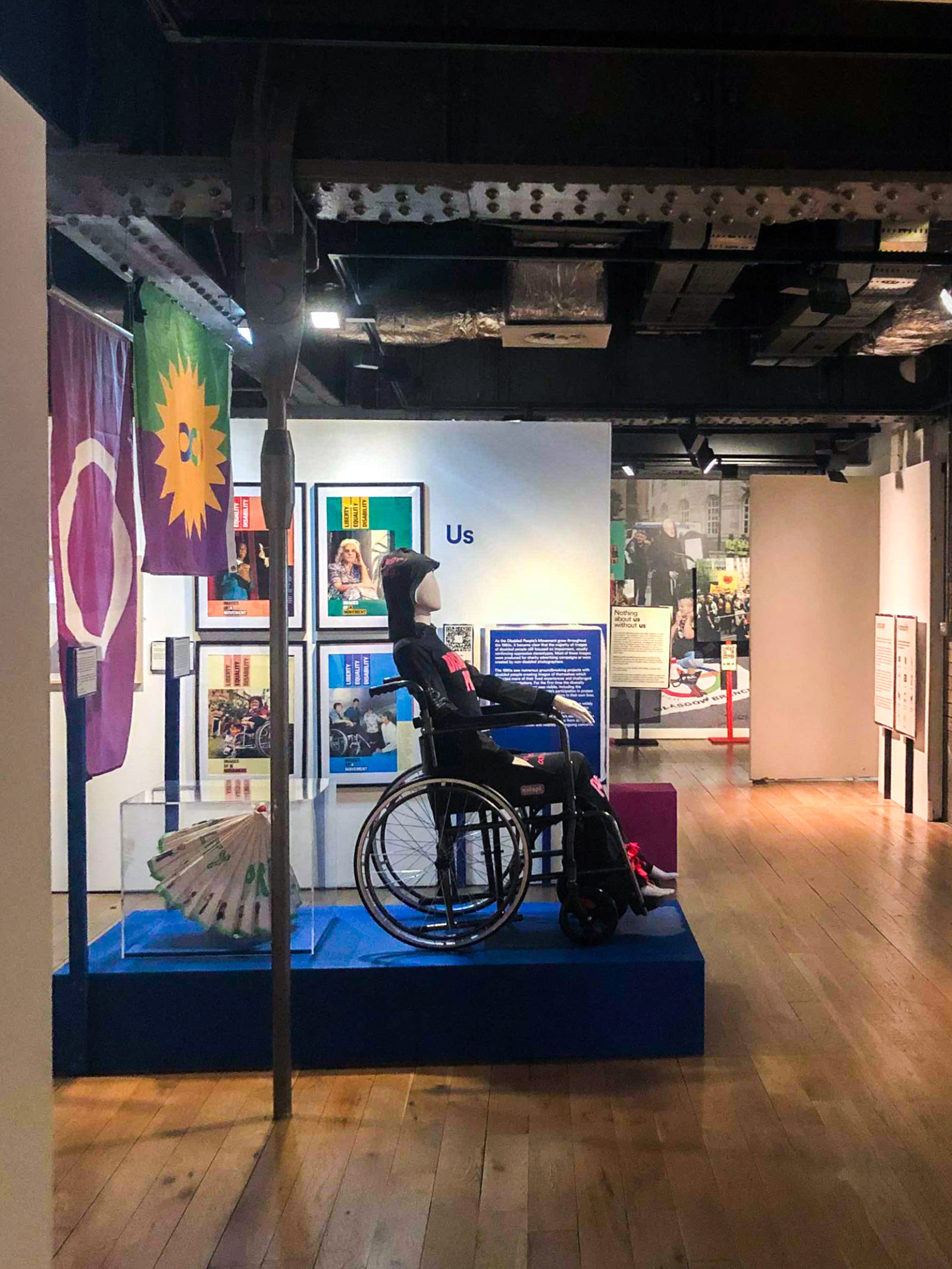 Photograph of the exhibition in people's history museum, features a mannequin in a wheelchair with flags and framed posters in the background.
