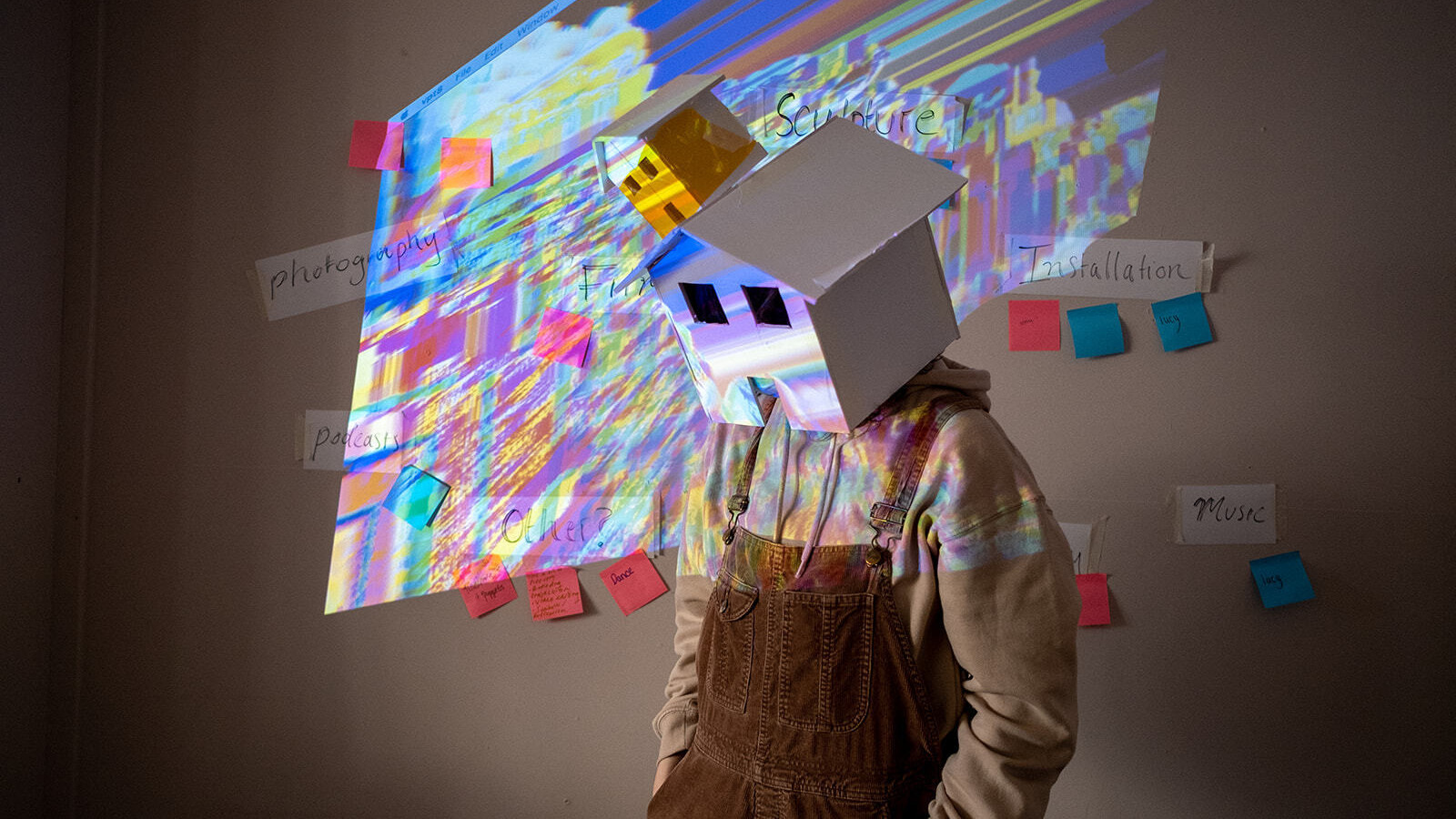 A person in dungarees wears a house-shaped mask on their head. A multi-coloured light projects onto the mask
