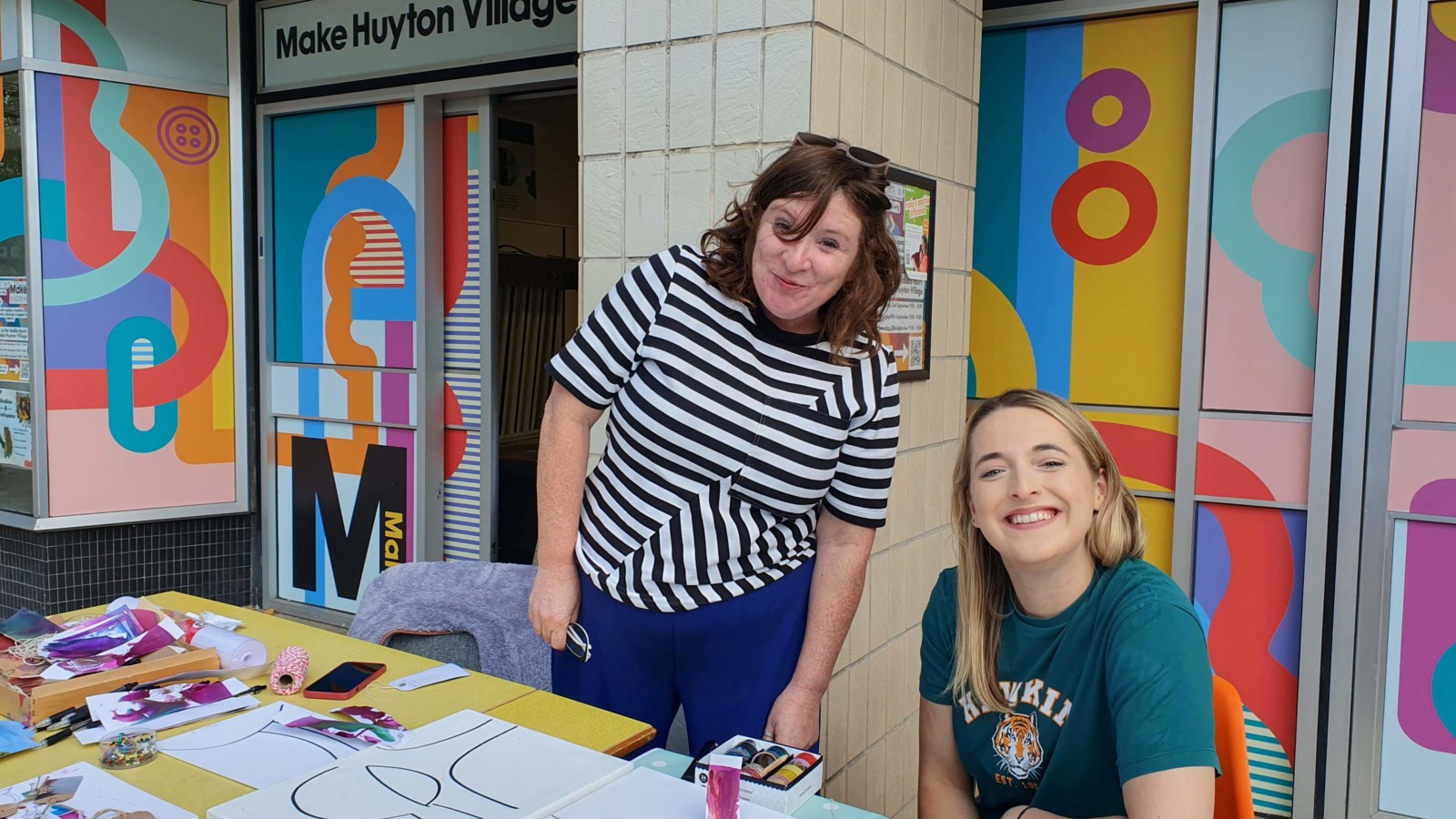 Producer Rhyannon and Heart of Glass collaborator Cathy Cross sit at a table outside the front of Make Huyton Village smiling at the camera. There are craft materials and heart of glass flyers on the table.