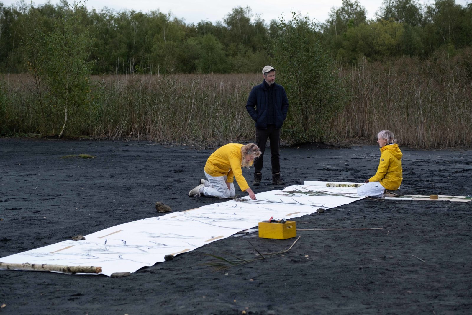 Kerry and Emma are knelt beside the long roll of paper, Kerry is drawing and Emma is talking to a man who is stood up beside them. Kerry and Emma are both wearing white overalls and yellow raincoats.