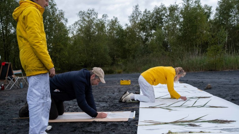 Traces Impressions and Digressions by Kerry Morrison is photographed taking place on the Slag heap at Colliers moss, Kerry is placing stalks on a large roll of paper and a man is doing a charcoal rubbing of of a stalk.