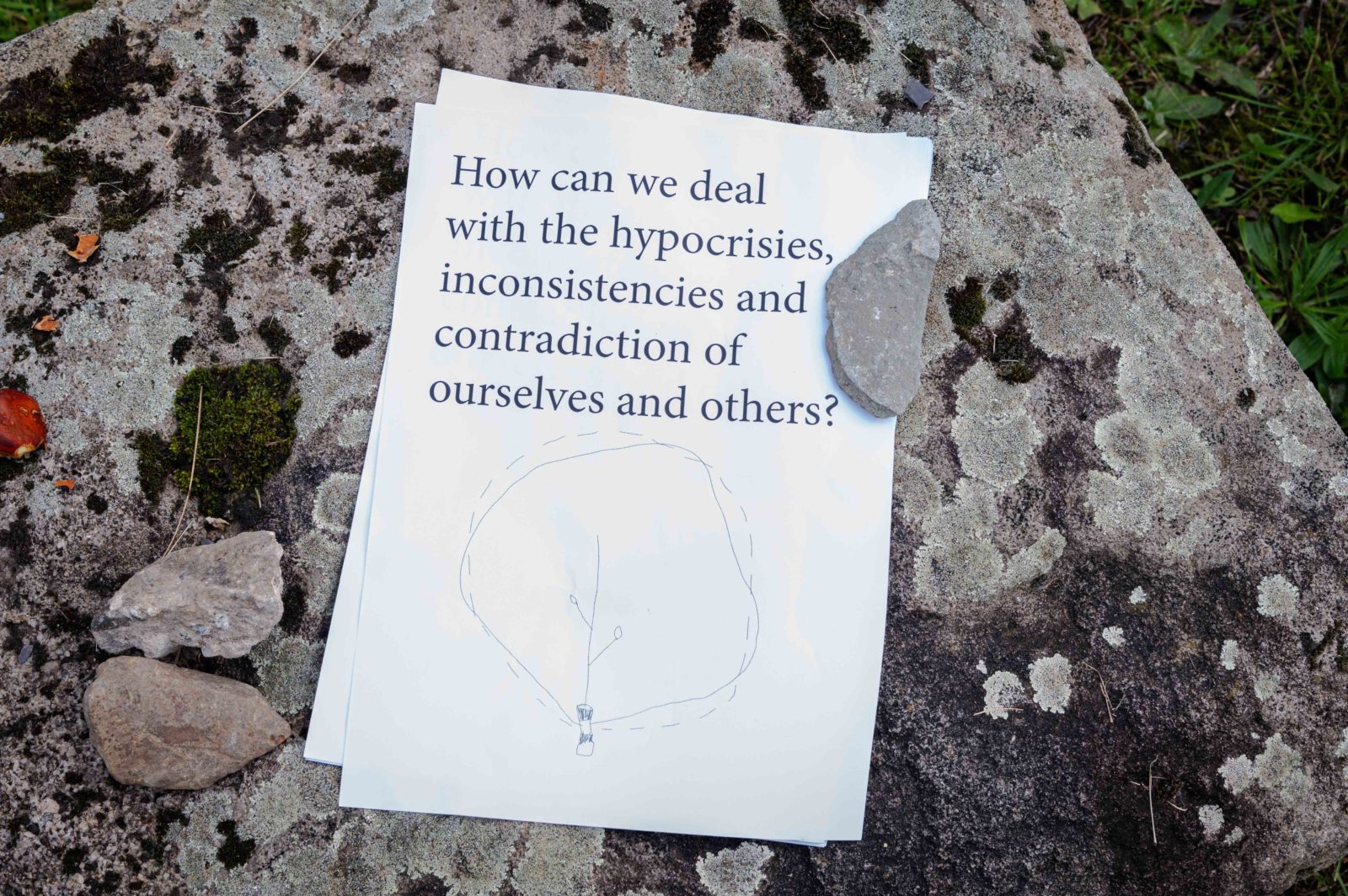 A sheet of paper that reads 'How can we deal with the hypocrisies, inconsistencies and contradiction of ourselves and others?', with a simple line illustration underneath is photographed on a rock.