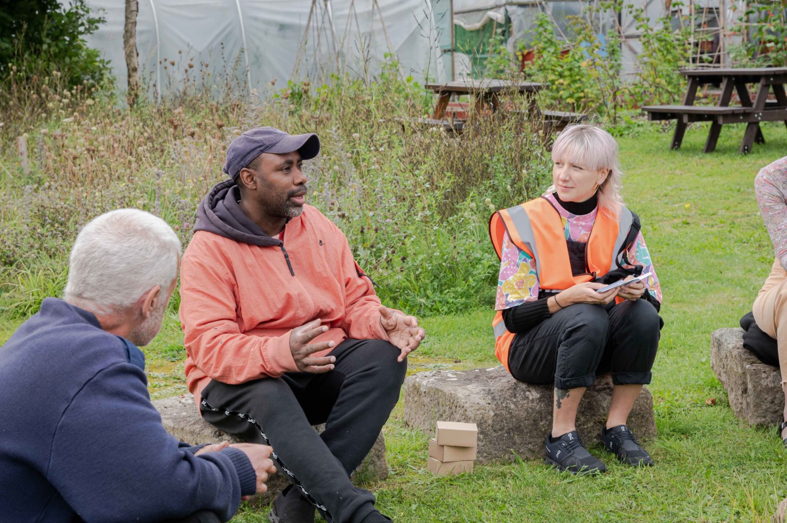 Harun Morrison is photographed speaking at the workshop he is leading. He is sat with a group of people stood outdoors on a circle of rocks.