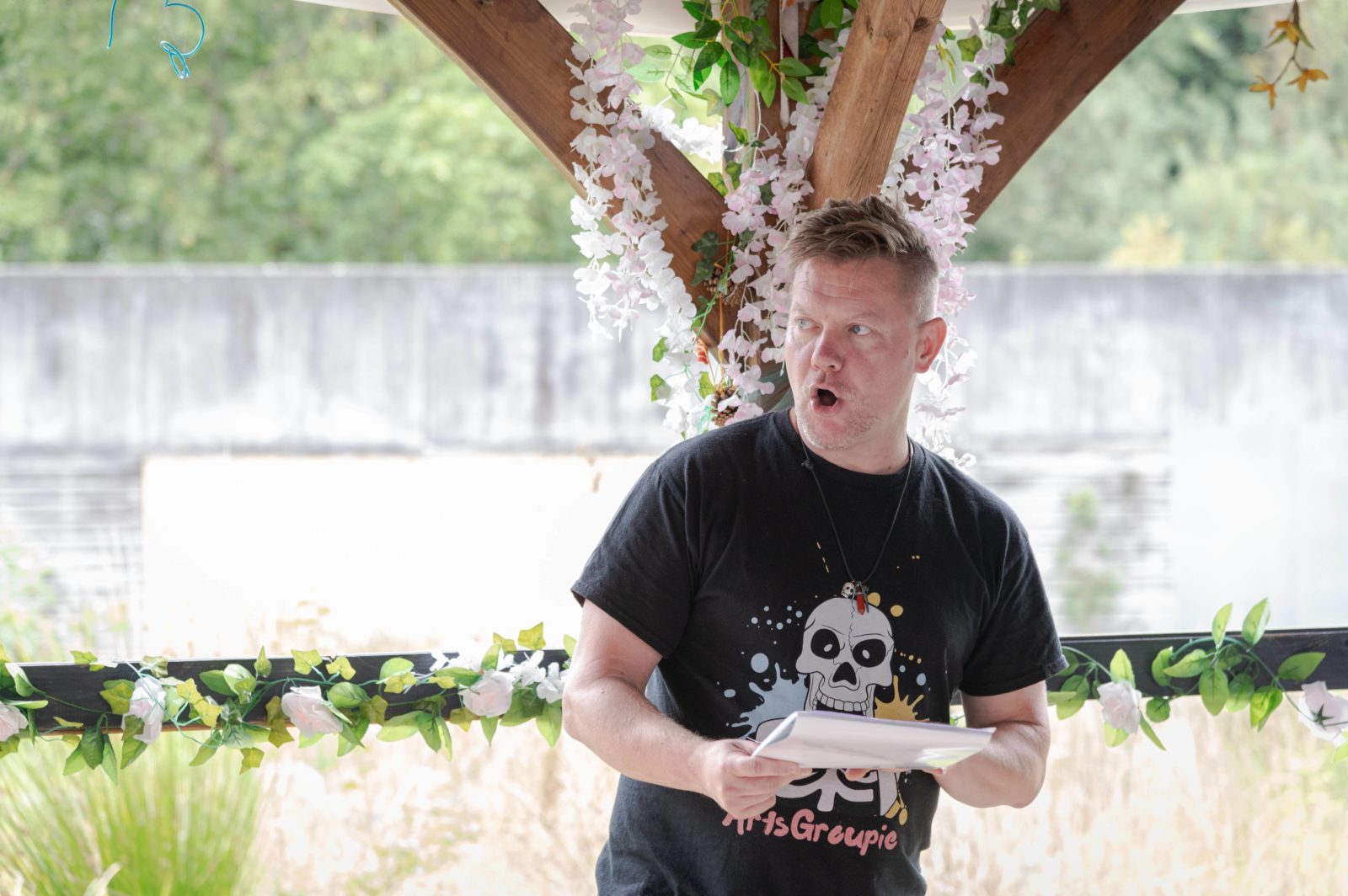 John Maguire is reading poetry off a sheet of paper in front of a wooden pillar with flowers hanging from it.