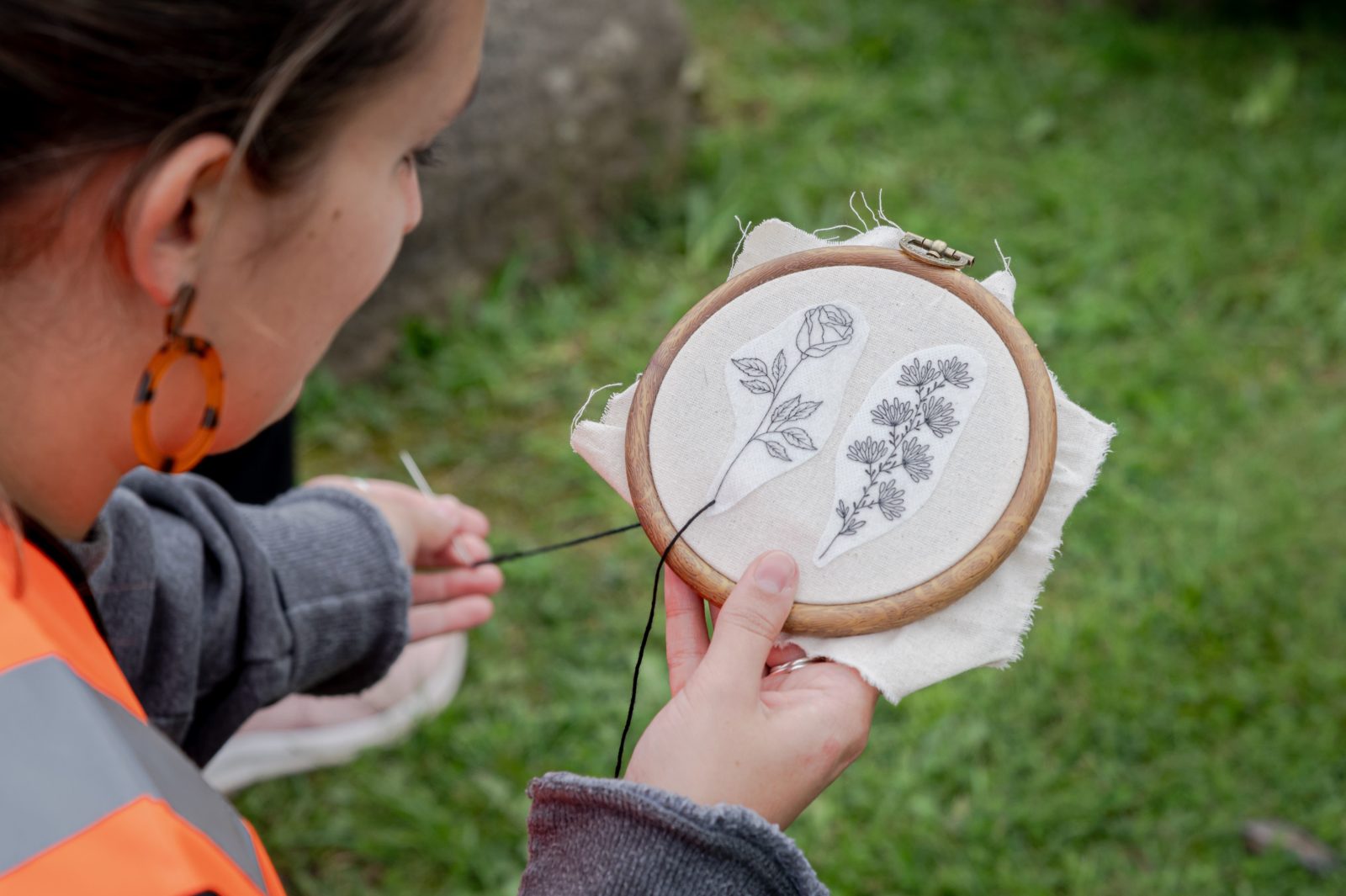 A girl is photographed embroidering 2 flowers onto an embroidery hoop outdoors.