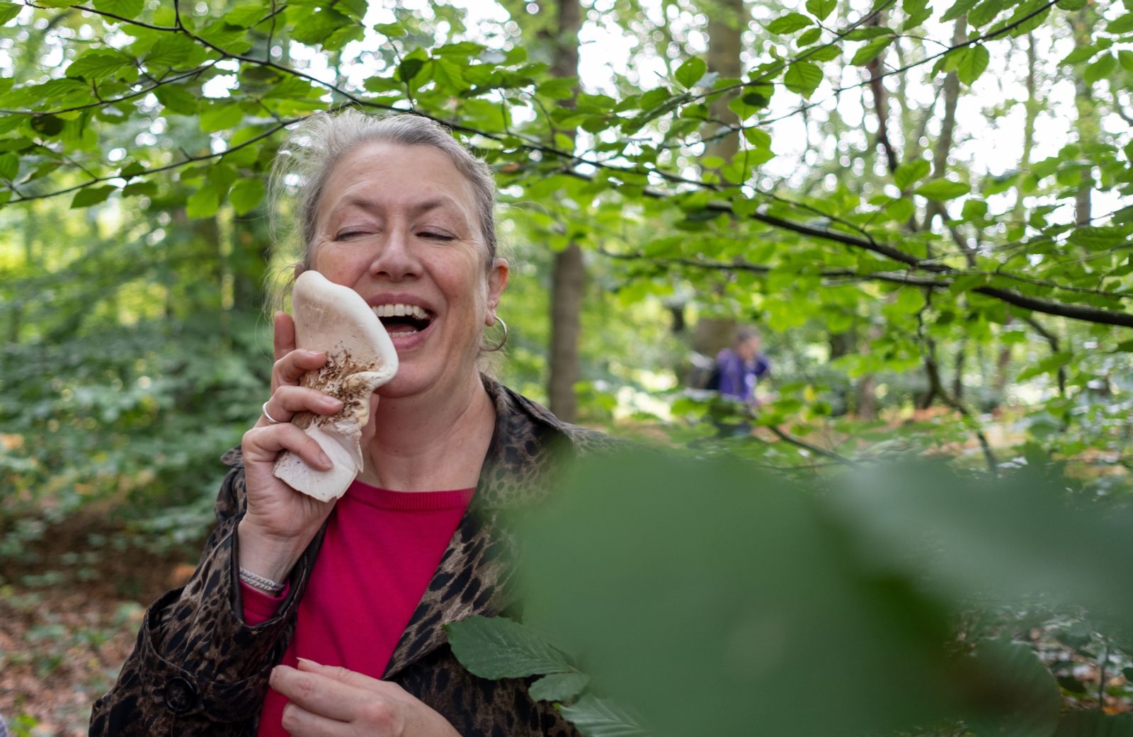 Jane Lawson is photographed holding a large flat mushroom up to her cheek in Stanley Bank Wood, she is laughing.