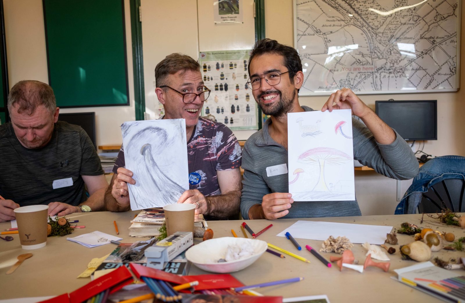 Two people sat at a table in the environment centre hold up their drawings of mushrooms for the camera whilst smiling.