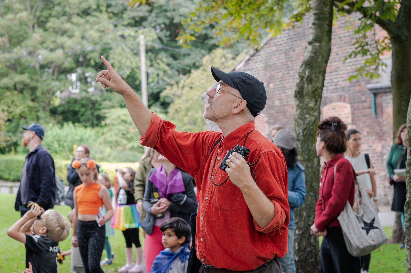 Paul is photographed at the Avian Able. He is looking up towards the sky and pointing. There are families in the background looking around in the same direction as hime, a little boy look through binoculars.